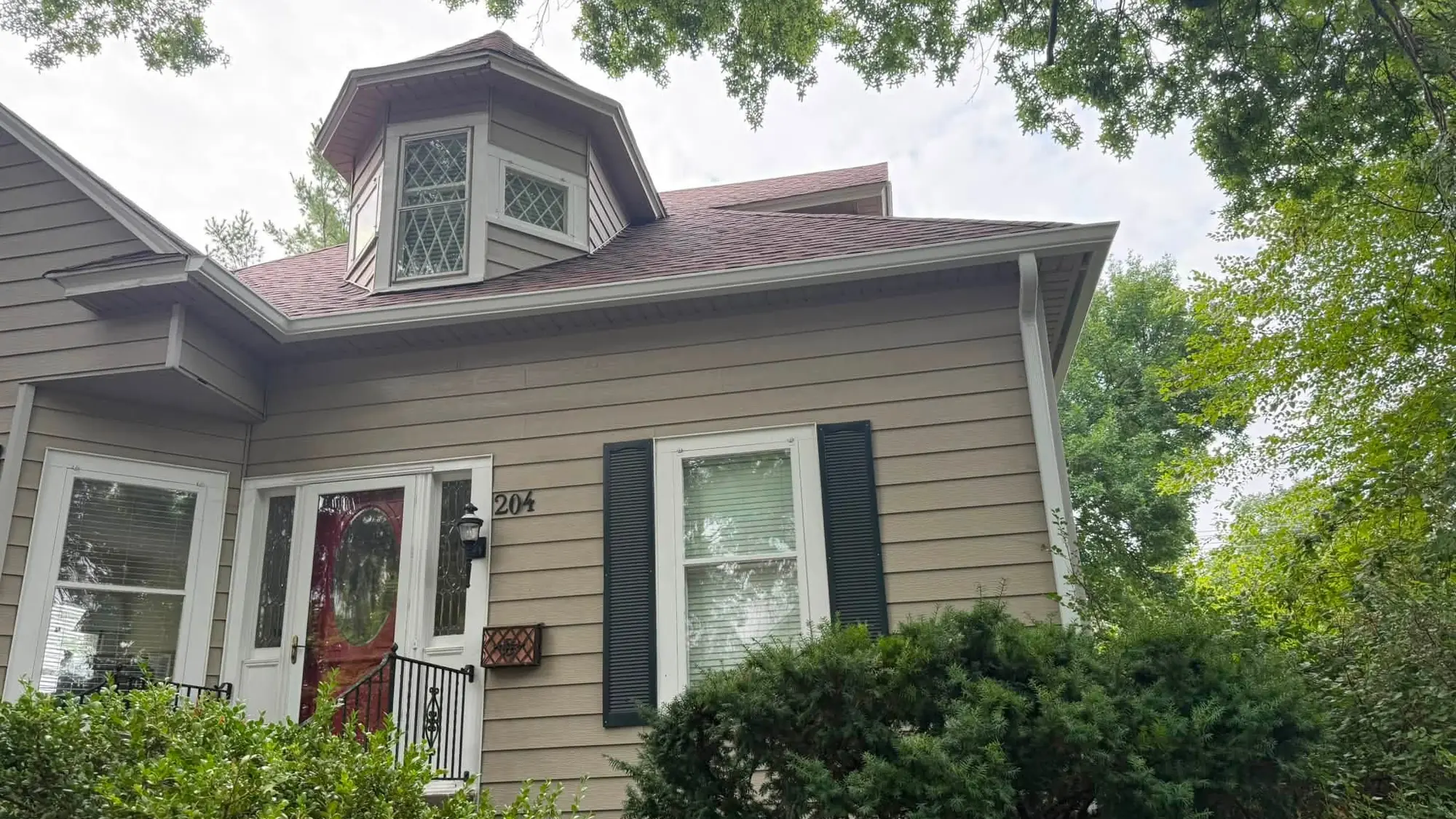 Tan bungalow with a bay dormer and white gutter, surrounded by mature trees
