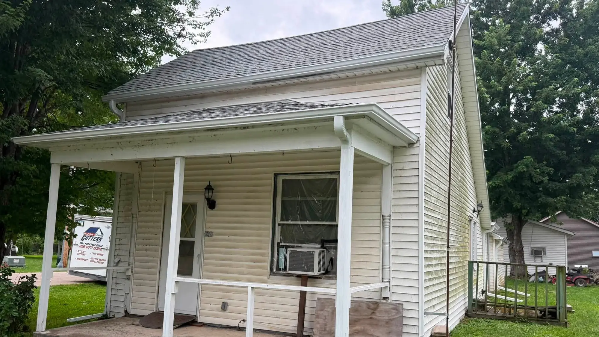 Older white clapboard cottage with a covered porch and new white gutter, the company yard sign staked in the grass
