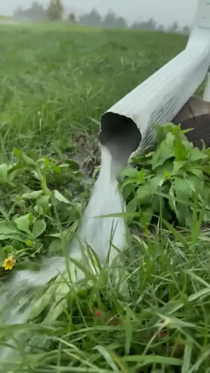 White downspout discharging a heavy stream of rainwater onto grass away from a building