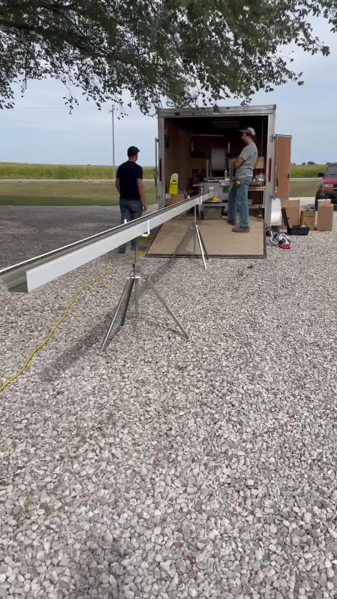 A full length of newly formed seamless gutter resting on roller stands beside the open company trailer, cornfield behind