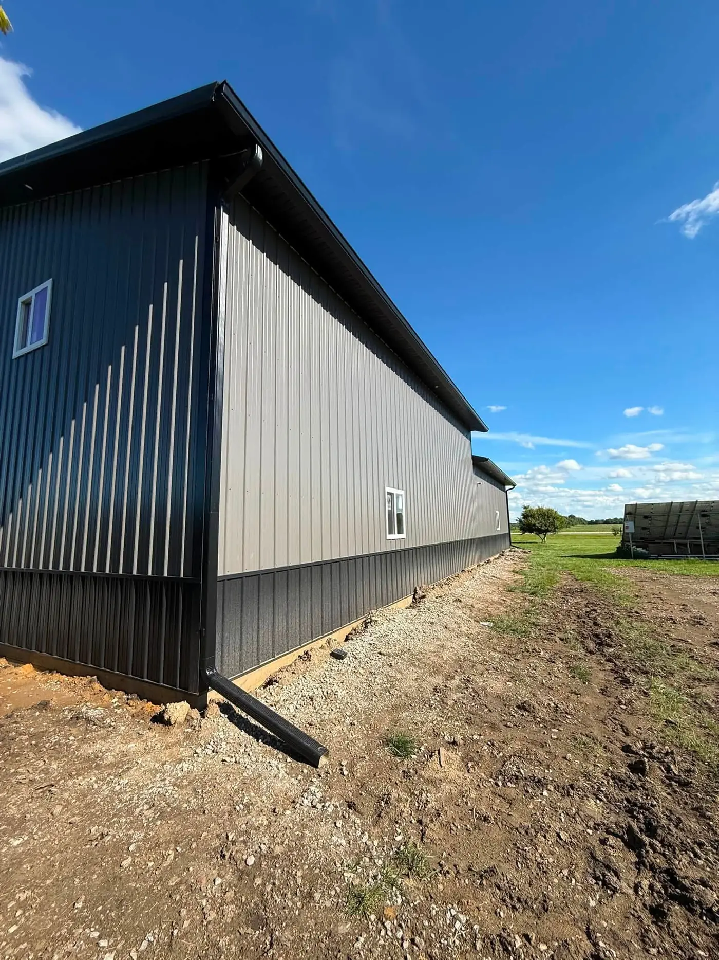 Long black and gray pole barn with a black gutter and downspout to fresh gravel