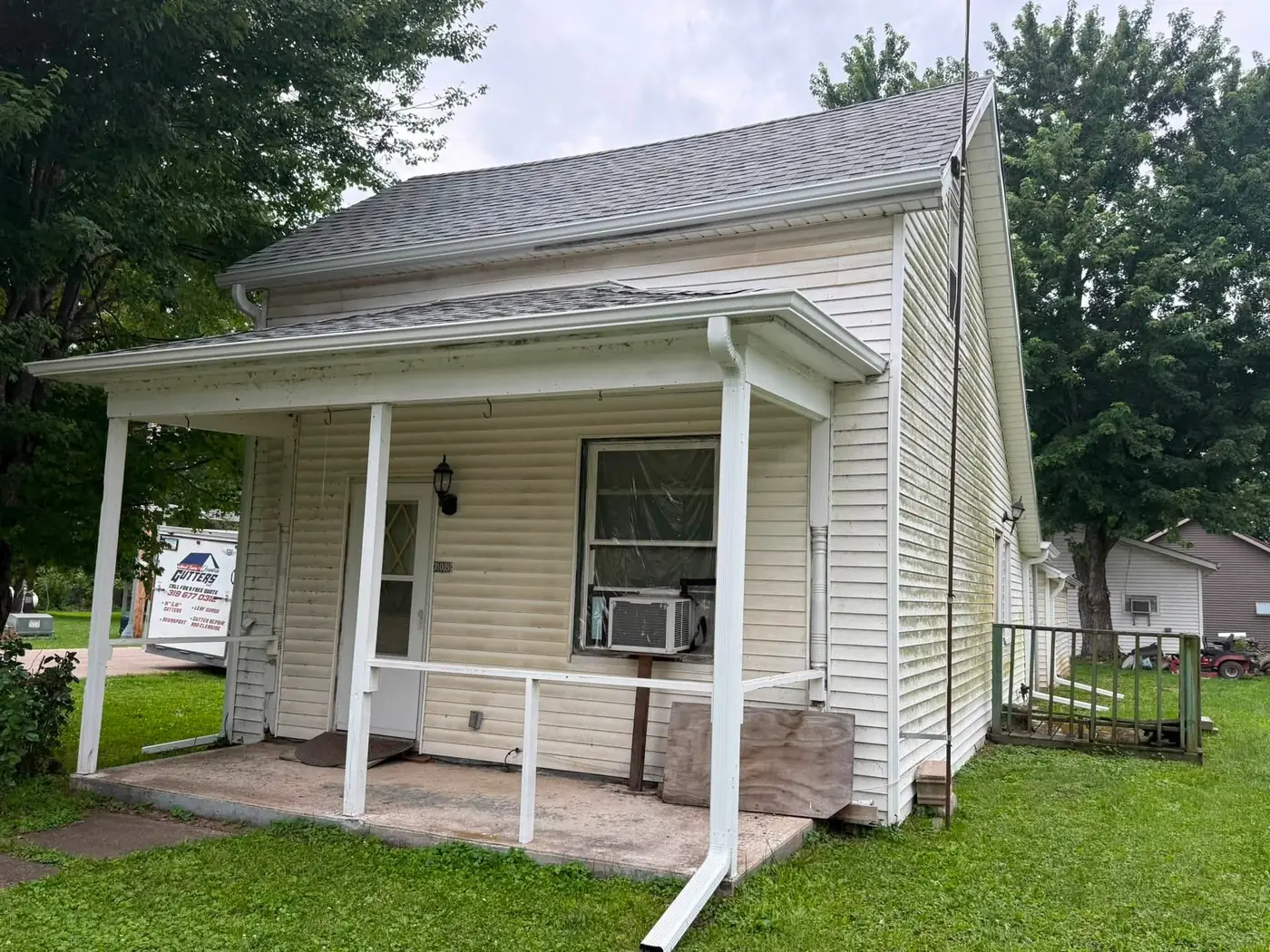 Older white clapboard cottage with a covered porch and new gutter, company yard sign in the grass