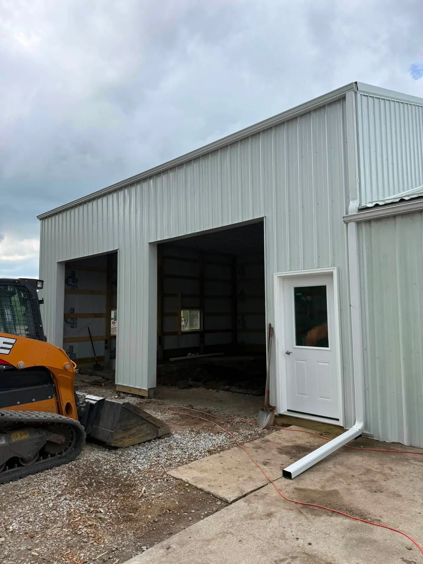 White metal shop still under construction with a skid steer and a downspout section on the concrete